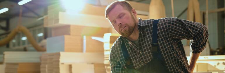 man in storage house full of boxes working bent over with hand on back in pain with face scrunched up